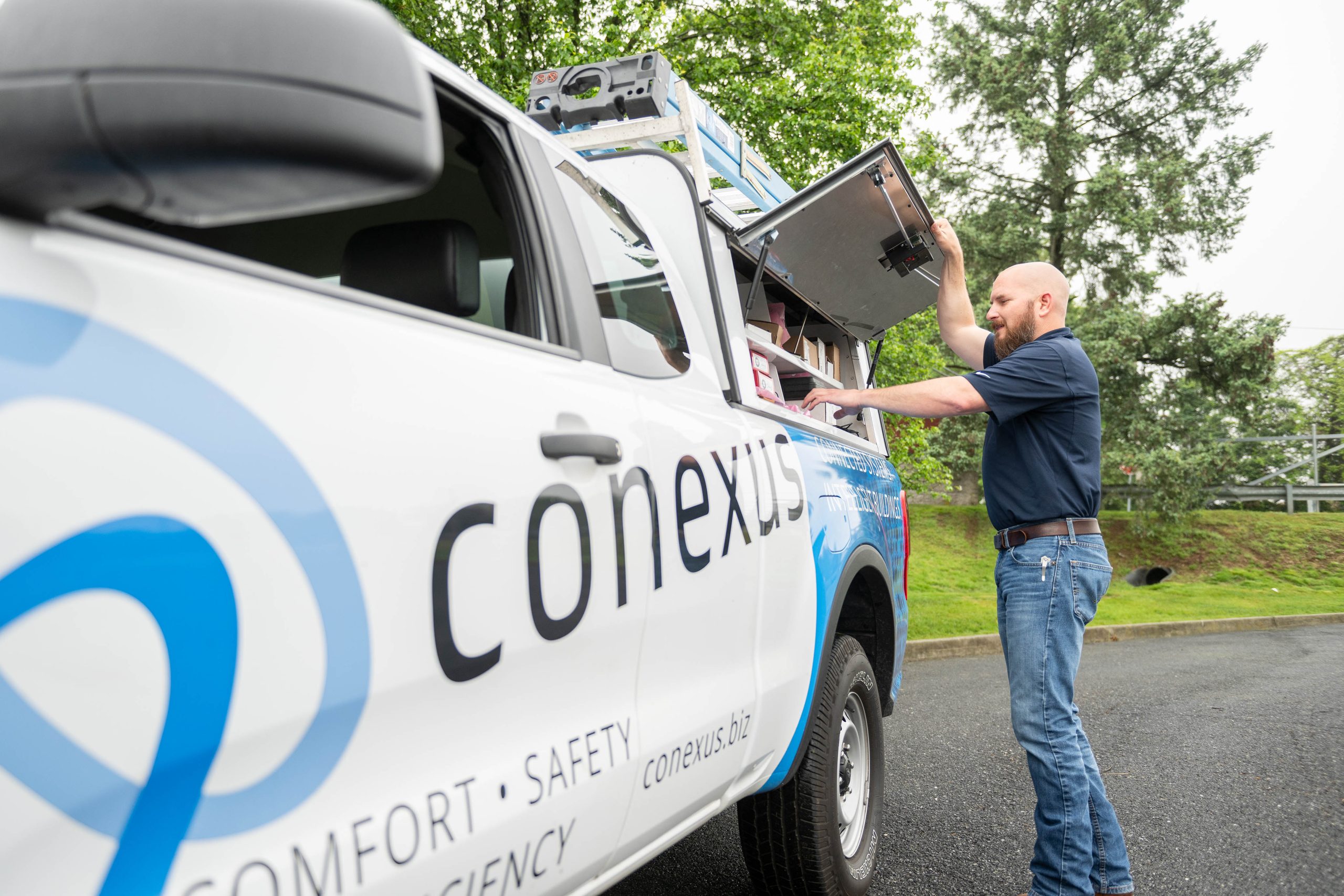A Conexus Service Technician with a bald head and beard is seen standing by his Conexus truck. The side panel is open as he grabs tools from inside to provide top-notch service to his customer.